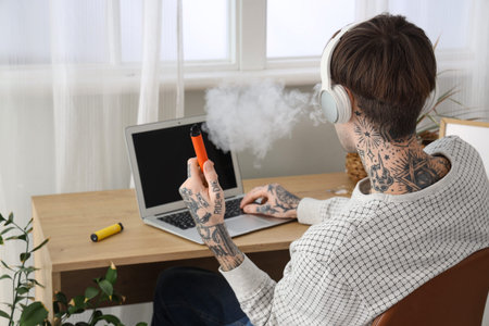 Young man in headphones smoking disposable electronic cigarette and using laptop on table at home,...