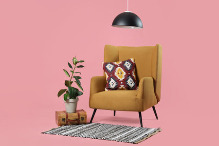 Comfortable armchair with cushion and plant on suitcase in pink background
