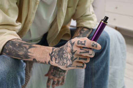 Young tattooed man with disposable electronic cigarette sitting on bed at home, closeupの写真素材