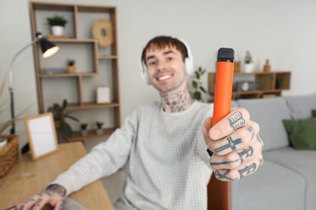 Young man in headphones smoking disposable electronic cigarette and using laptop on table at home