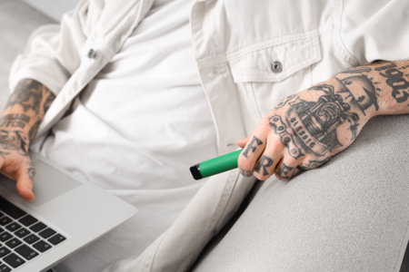 Young man with disposable electronic cigarette and laptop at home, closeupの写真素材