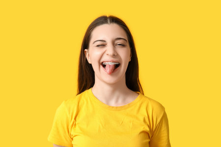 Happy young woman in stylish yellow t-shirt showing tongue on color background
