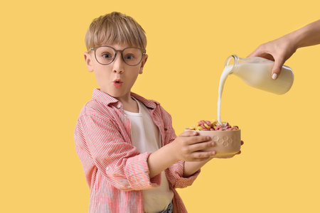 Surprised little boy with bowl of cereal and hand with bottle of milk on yellow backgroundの写真素材
