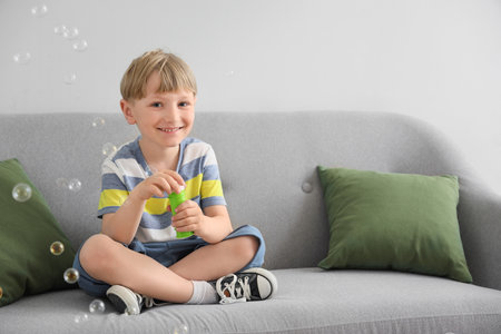 Cute little boy sitting on sofa and blowing soap bubbles at homeの写真素材