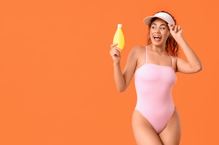 Happy young African-American woman in swimsuit with bottle of sunscreen cream on orange backgroundの写真素材
