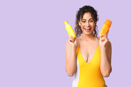 Happy young African-American woman in swimsuit with bottles of sunscreen cream on lilac background