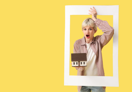 Portrait of surprised young woman with house model and photo frame on yellow background. Mortgage...