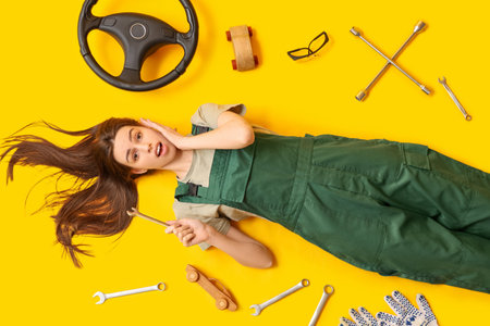 Surprised young female auto mechanic with different tools and steering wheel lying on yellow backgroundの写真素材