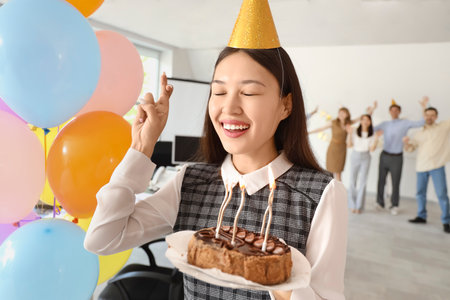Young Asian businesswoman with cake making wish at birthday party in officeの写真素材