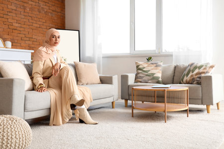 Stylish Muslim woman in hijab sitting on sofa at homeの写真素材