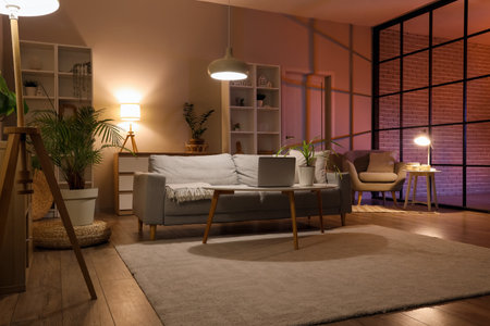 Sofa, table with modern laptop and glowing lamps in interior of living roomの写真素材