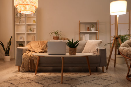 Stylish interior of living room with sofa, armchair, coffee table with laptop and glowing lampsの写真素材
