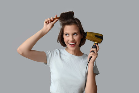 Beautiful young happy woman with bob hairstyle holding dryer and brush on gray background