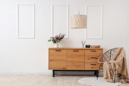 Vase with lilacs on wooden chest of drawers, lamp and armchair in stylish roomの写真素材
