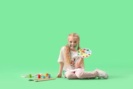 Cute little girl in paint with palette sitting on green backgroundの写真素材