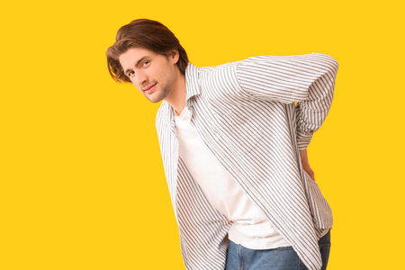 Handsome young man suffering from back pain on yellow background