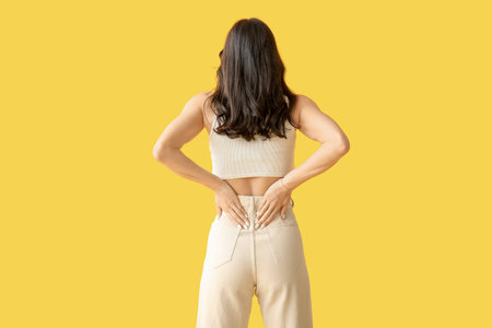 Beautiful young woman suffering from back pain on yellow background