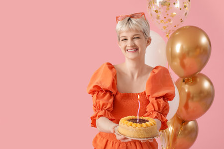 Happy young woman holding birthday cake with burning candles and balloons on pink backgroundの写真素材