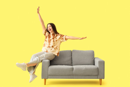 Happy young woman sitting on gray sofa against yellow backgroundの写真素材