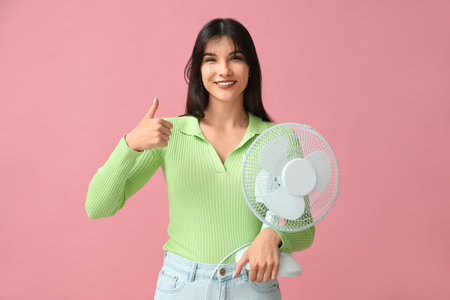 Young woman with electric fan showing thumb-up on pink backgroundの写真素材