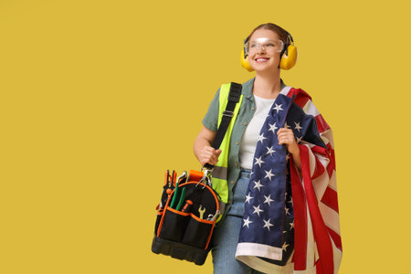 Young female carpenter with USA flag and tool bag on yellow background. Labor Day celebration