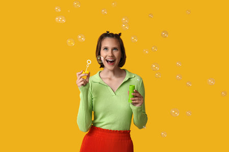 Happy teenage girl blowing soap bubbles on yellow background