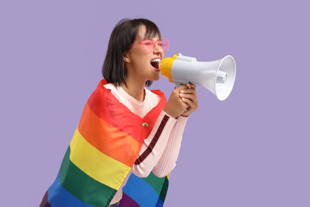 Beautiful young woman with megaphone and LGBT flag on lilac backgroundの写真素材