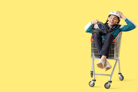 Sporty young man with steering wheel in shopping cart racing on yellow background