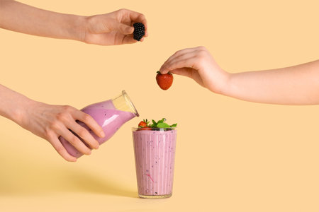 Woman pouring fresh smoothie from bottle into glass and female hands with berries on yellow backgroundの写真素材