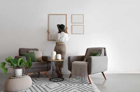 Young woman hanging big blank frame on light wall at home, back view