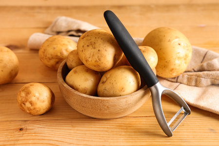 Bowl with raw potatoes and peeler on wooden backgroundの写真素材