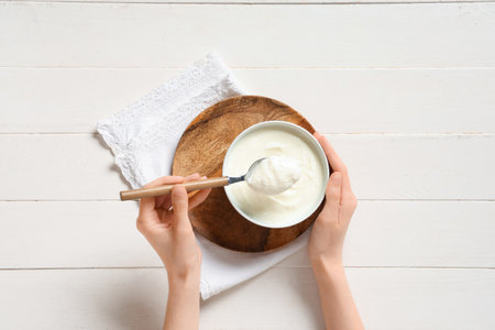 Female hands with bowl of tasty yogurt and spoon on white wooden backgroundの写真素材