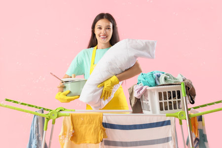 Overwhelmed Asian mother with baby, cooking pot, soap bubbles and clothes on dryer against pink background. Multitasking conceptの写真素材