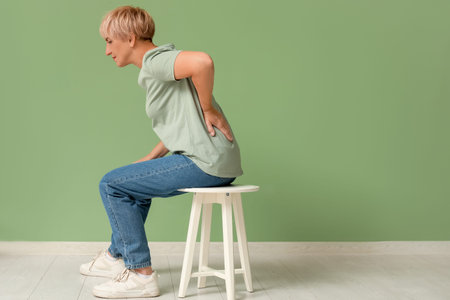 Mature woman suffering from back pain while sitting on chair on green background