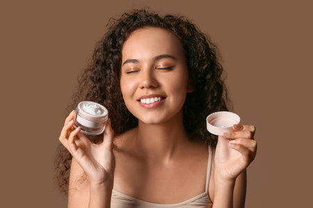 Beautiful young African-American woman with jar of face cream on brown backgroundの写真素材