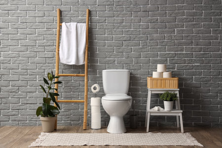 Interior of restroom with toilet bowl, ladder and shelving unit with toilet paper near gray brick wallの写真素材