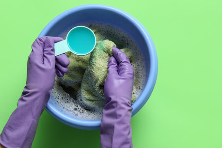 Woman in rubber gloves adding laundry detergent to clothes in plastic basin on green backgroundの写真素材