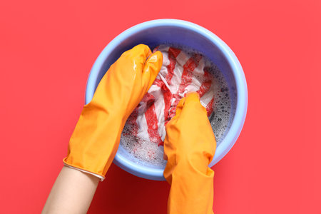 Woman in rubber gloves washing clothes in plastic basin on red background