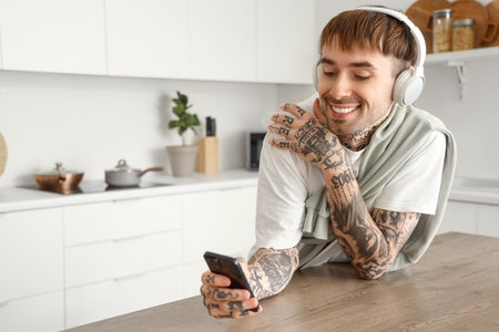 Young tattooed man with headphones and mobile phone listening to music at table in kitchenの写真素材