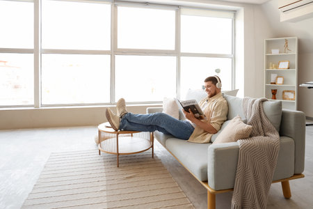 Young bearded man in headphones reading magazine on sofa at homeの写真素材