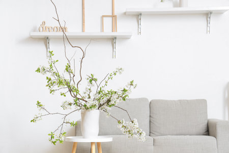 Vase with blooming branches on coffee table near cozy sofa in white living roomの写真素材