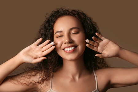 Beautiful young African-American woman applying cream on face against brown backgroundの写真素材