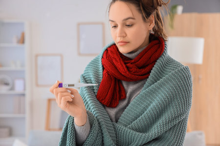 Sick young woman with thermometer and blanket at home