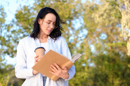 Pretty young woman with cup of coffee reading book in park
