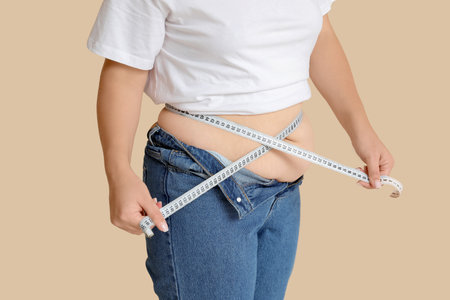 Overweight woman in tight jeans measuring her belly on beige background. Excess weight conceptの写真素材