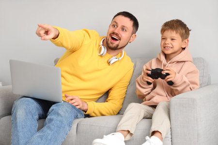 Happy little son playing video games and his father working with laptop on sofa at home