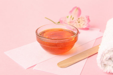 Glass bowl with sugaring paste, wax strips, wooden stick and flower on pink background, closeupの写真素材