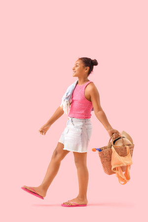 Happy African-American girl with wicker bag and beach accessories on pink backgroundの写真素材