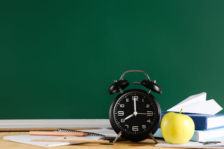 Apple, alarm clock, books and paper plane on desk near chalkboard. Back to school concept