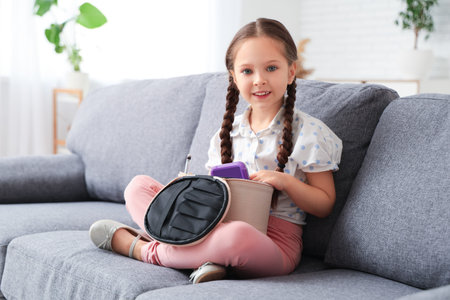 Cute little girl with cosmetic bag sitting on sofa at homeの写真素材